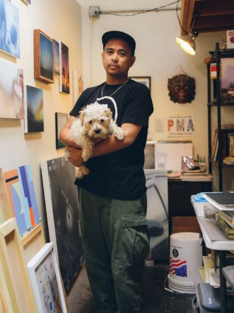 Adult male artist in a black t-shirt and olive-green pants holding a small dog in an art studio surrounded by paintings and art supplies.