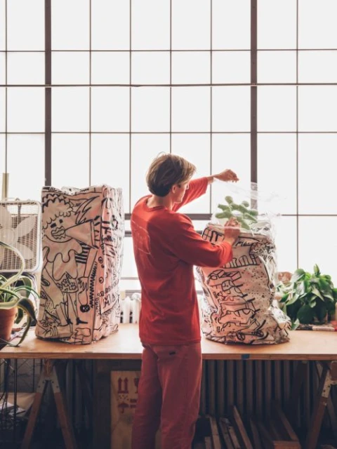 Person in a red jumpsuit tending to a potted plant, next to a large hand-drawn sack in a bright room with large windows.