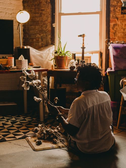 Adult female arranges cotton plants on a wooden floor, indoors. Natural light streams through a window in the background.