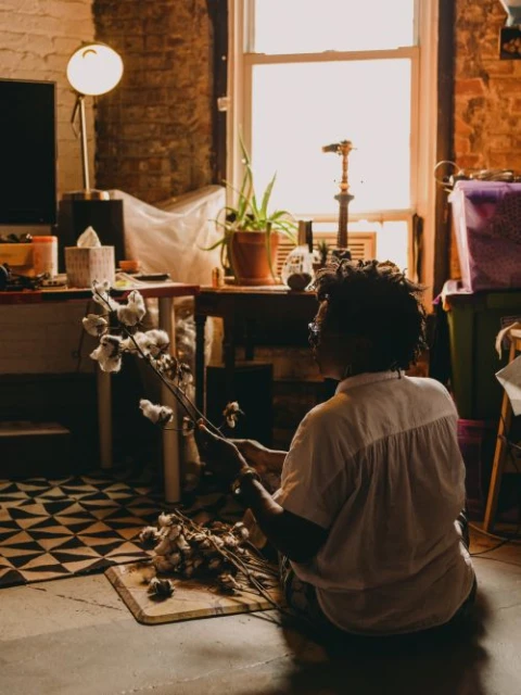 Adult female arranges cotton plants on a wooden floor, indoors. Natural light streams through a window in the background.