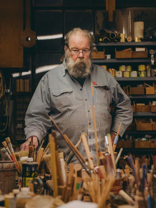 Older male woodworker in a striped shirt, standing in his workshop among paint brushes and woodworking tools with soft, warm lighting.