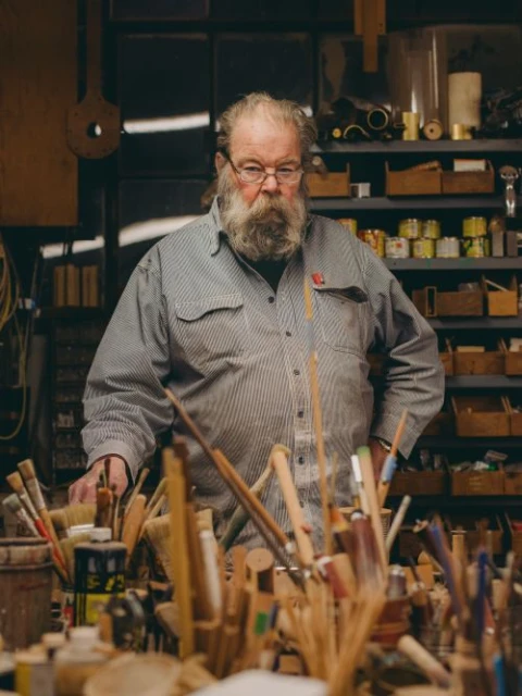 Older male woodworker in a striped shirt, standing in his workshop among paint brushes and woodworking tools with soft, warm lighting.