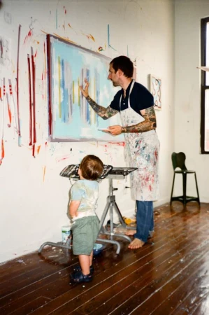 An adult male artist and a young child stand in front of a vibrant painting in a paint-splattered studio, working on the canvas together.