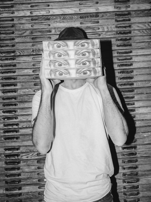 Person holding four boxes with eye illustrations in a black and white photo. Man in hat and white shirt stands in front of drawers.