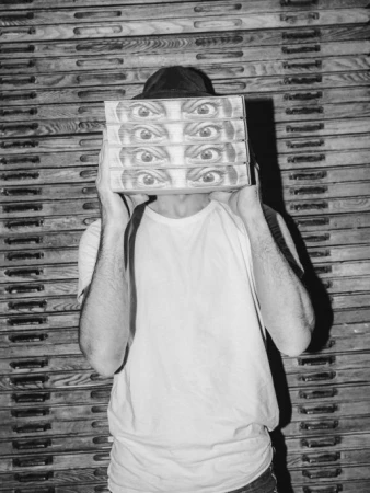 Person holding four boxes with eye illustrations in a black and white photo. Man in hat and white shirt stands in front of drawers.