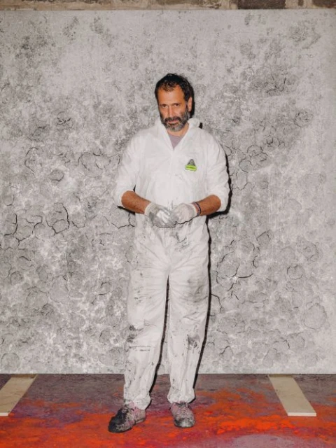 Full-length shot of a man in a white jumpsuit and gloves standing against a textured gray and white wall with a red and orange floor.