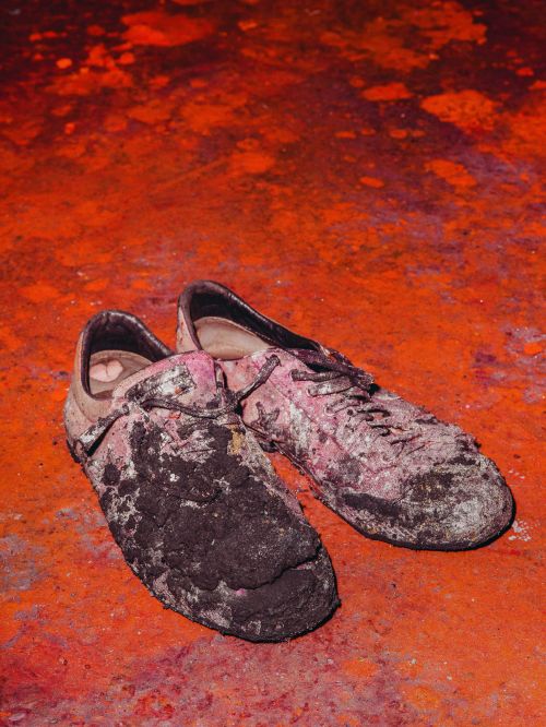 Pair of dirty athletic shoes with laces on a red mottled surface, possibly clay, suggesting post-game or exercise activity with signs of wear.