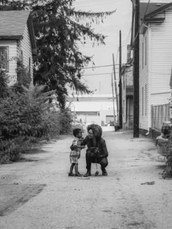 Monochrome street-level photograph of a woman kneeling to talk with a young child in an urban alleyway.