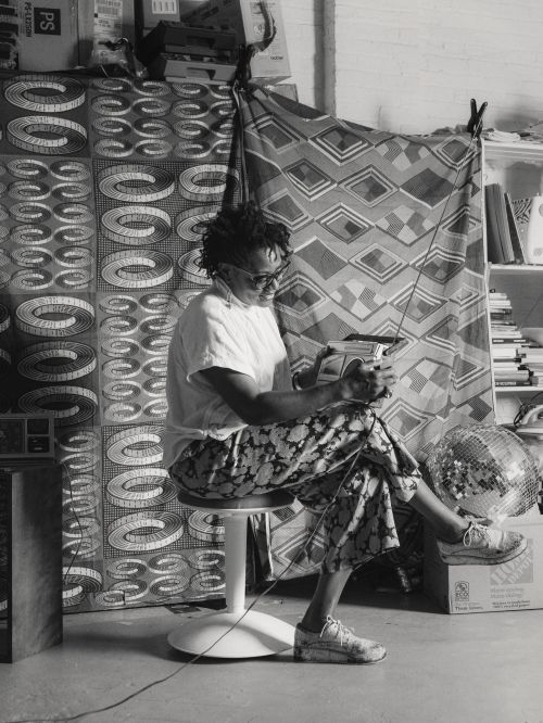 Black and white portrait of a person sitting on a stool, holding a handheld device, in a room with patterned fabrics in the background.