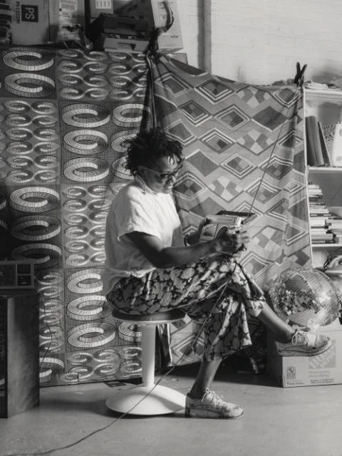 Black and white portrait of a person sitting on a stool, holding a handheld device, in a room with patterned fabrics in the background.