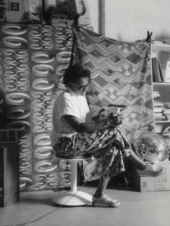 Black and white portrait of a person sitting on a stool, holding a handheld device, in a room with patterned fabrics in the background.