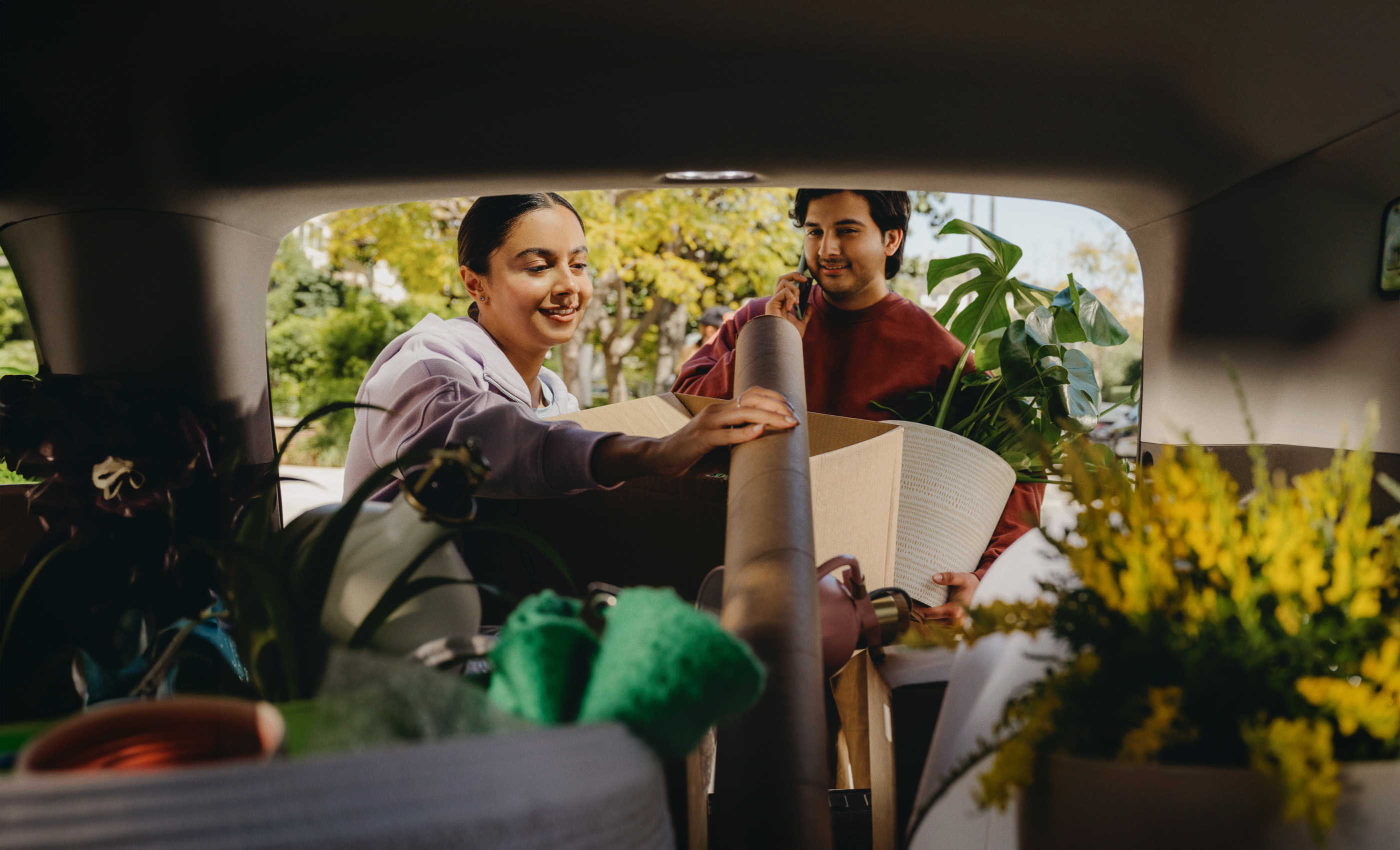 Couple moving with boxes and plants in car. Young woman and man loading cardboard containers and potted greenery into vehicle trunk outdoors.
