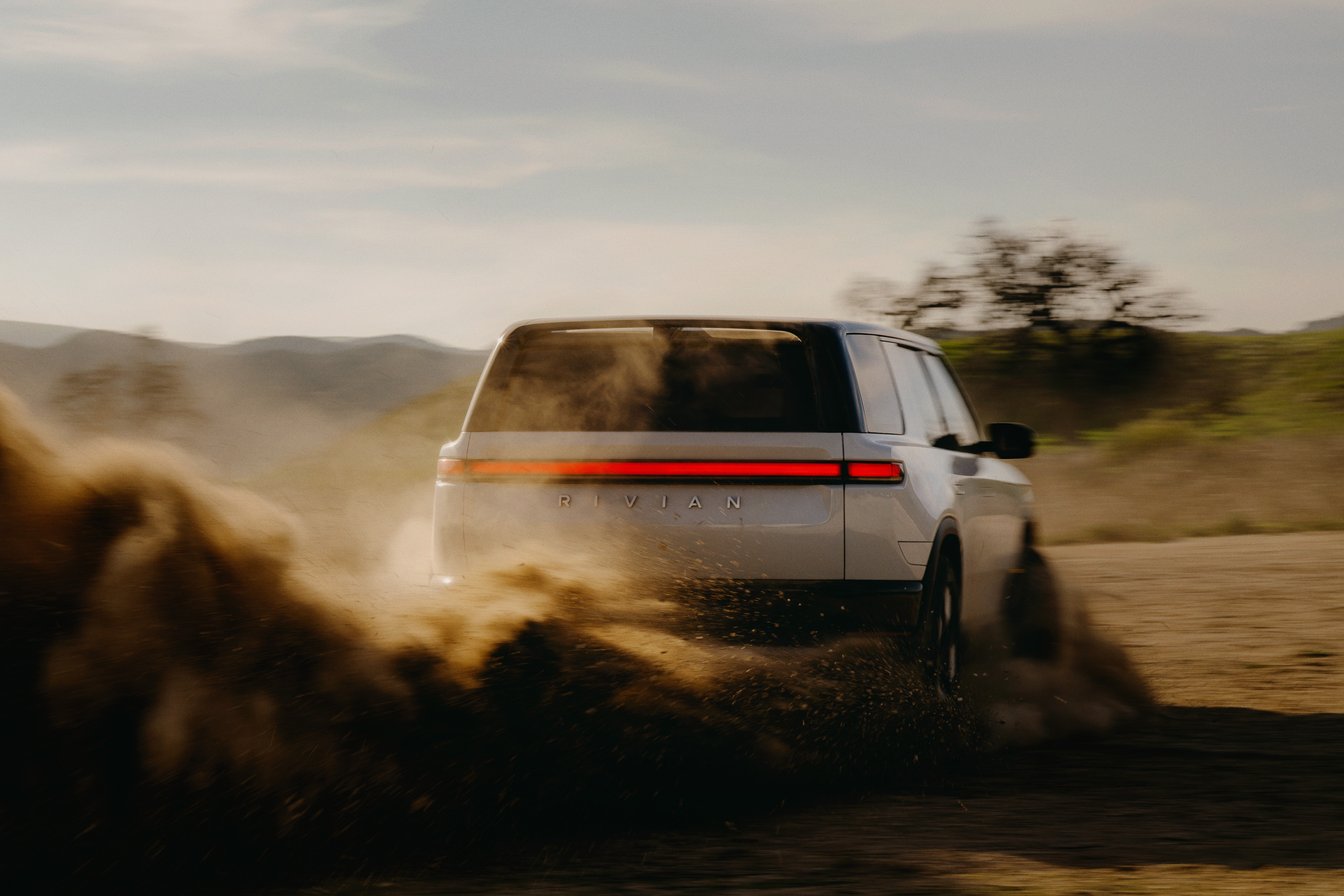Rivian SUV driving through a dusty, rural landscape. The vehicle's rear is visible, with a backdrop of mountains and open fields.
