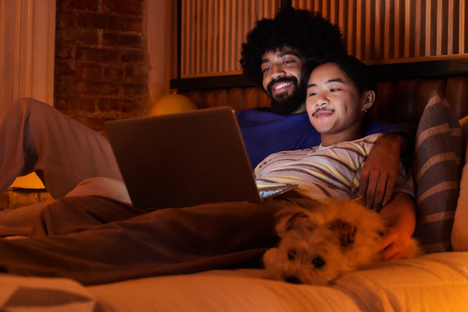Two men relaxing on a bed together with a small dog while looking at an open laptop in a warmly lit room.