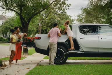 Family unloading potted flowers from the bed of a silver pickup truck parked on a residential suburban street