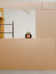 Man wearing a bucket hat peeking over a large cardboard sheet in a studio with scaffolding visible