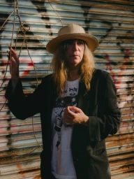 Portrait of a person wearing a beige hat and dark jacket against a corrugated metal shutter backdrop.