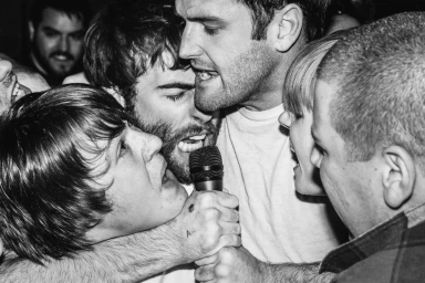 Monochrome close-up photograph of a crowd of young adults singing passionately into a handheld microphone in a tight group.
