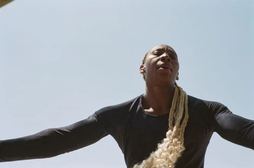 Adult with blonde braided hair wearing a black top is standing outside against a blue sky in a low angle full shot.