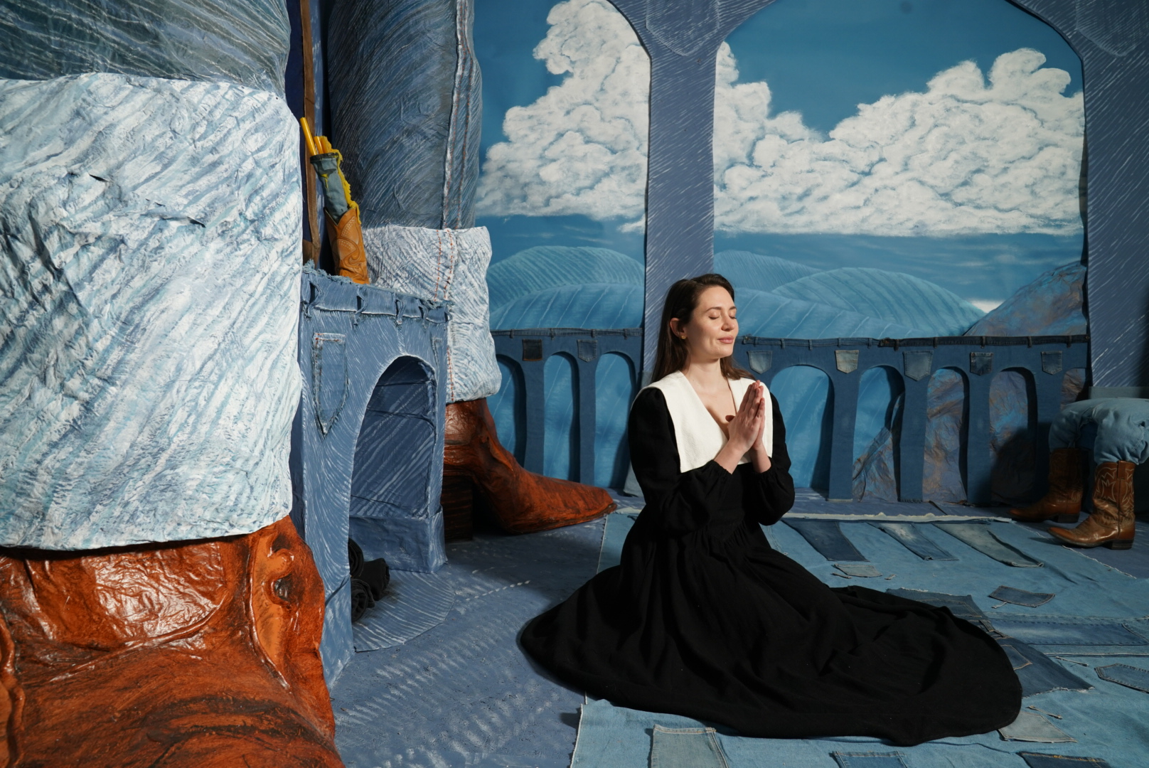 Woman kneels with hands clasped in an expressive stage environment featuring denim patchwork, painted mountains, and exaggerated features.