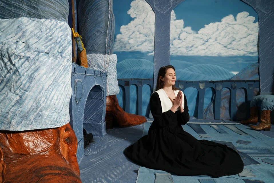 Woman kneels with hands clasped in an expressive stage environment featuring denim patchwork, painted mountains, and exaggerated features.
