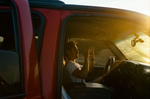 Man driving red SUV with open door in desert setting during daytime, bathed in sunlight, capturing a moment of travel and adventure.