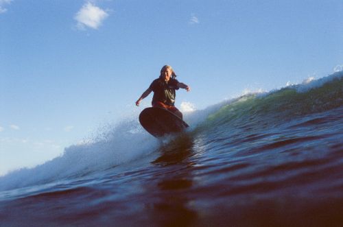 Adult surfer riding a wave on a surfboard against a blue sky. Water splashes around the surfboard as the surfer balances on the wave.