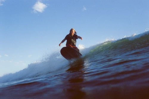 Adult surfer riding a wave on a surfboard against a blue sky. Water splashes around the surfboard as the surfer balances on the wave.