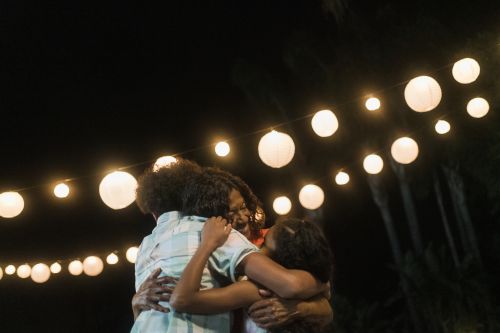 Three people, likely family, embracing outside at night under a string of glowing lanterns. Dark environment with some plants visible.