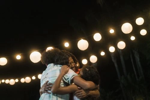 Three people, likely family, embracing outside at night under a string of glowing lanterns. Dark environment with some plants visible.
