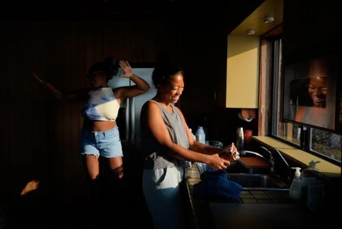 Two women in a sunlit kitchen. One African-American woman is washing dishes while another is jumping in the background.