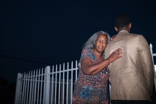 Elderly woman with grey hair affectionately touches the back of a young man in a tan jacket against a dark blue outdoor backdrop.