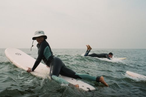 Two surfers in wetsuits on surfboards ride the ocean waves under an overcast sky, enjoying a day of watersports and recreation.