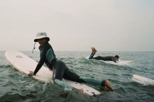 Two surfers in wetsuits on surfboards ride the ocean waves under an overcast sky, enjoying a day of watersports and recreation.