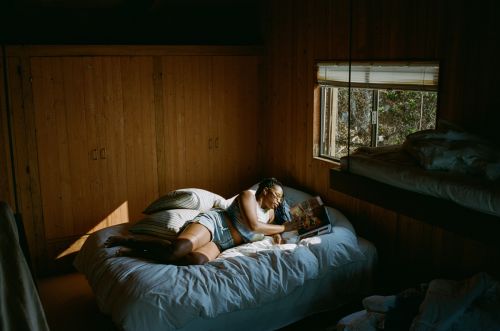 African-American woman relaxes in wood-paneled room, lounging on a bed reading a magazine by a window with a view to the outdoors.