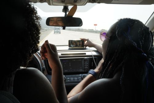 Two adult women in a car, one taking a picture with a smartphone. Road and other car visible through the windshield.