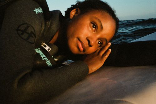 Portrait of a young adult female wearing a wetsuit, resting her head on her hand, in a marine environment with visible waves.