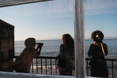 Three people near a reflective window overlooking the ocean on a sunny day. One person is taking a picture with a camera.