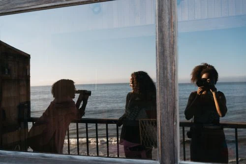 Three people near a reflective window overlooking the ocean on a sunny day. One person is taking a picture with a camera.