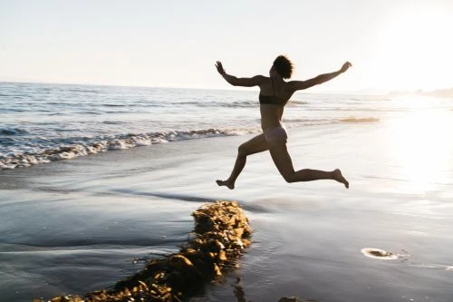 A woman jumps barefoot on a beach in a bikini. The scene is set at the ocean's edge, with the sun in the distance.