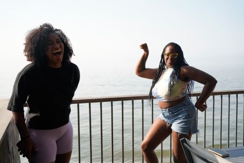 Two adult women posing outdoors on a deck with a water view: one laughing, one flexing arm muscles, wearing casual clothing and sunglasses.