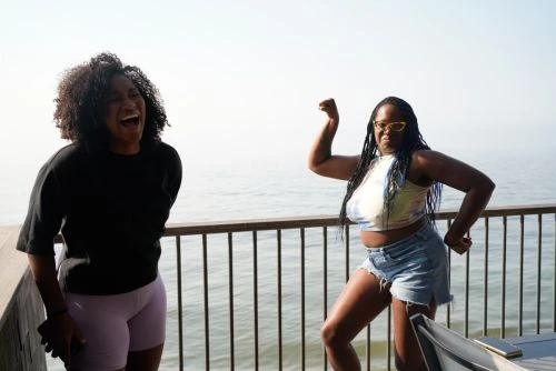 Two adult women posing outdoors on a deck with a water view: one laughing, one flexing arm muscles, wearing casual clothing and sunglasses.