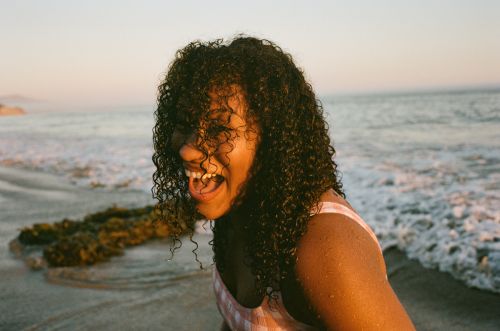 Adult female smiling on a beach at sunset with curly hair, wearing a patterned top, with the ocean and sky in the background.