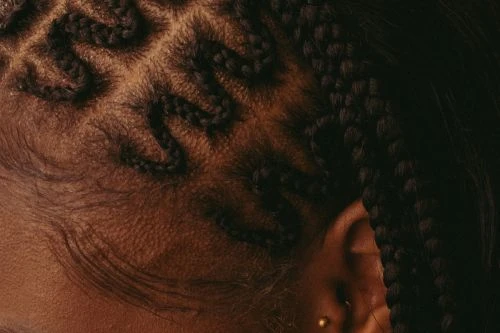 Close-up of textured dark skin with intricately braided hair in a wavy pattern near the ear, emphasizing detail and design.