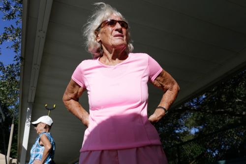 Elderly woman in pink sportswear stands confidently outside with another woman in the background on a sunny day.