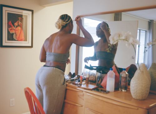 A woman applies makeup in front of a mirror in a brightly lit room with a dresser, a framed artwork, and white orchid blossoms.