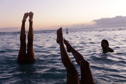 Three people are in the ocean, with two people's legs sticking straight up out of the water, and a third person swimming nearby.