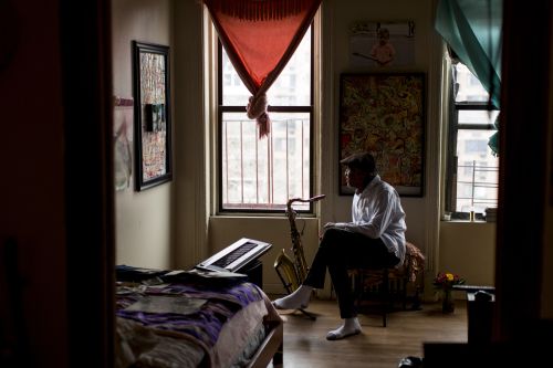 Man sitting indoors with musical instruments in a brightly lit room, framed by a doorway and featuring neutral and muted colors.