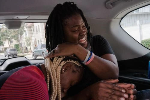 Two adult females embrace inside a car, one laughing and the other looking down with blonde braided hair. Buildings visible outside the window.