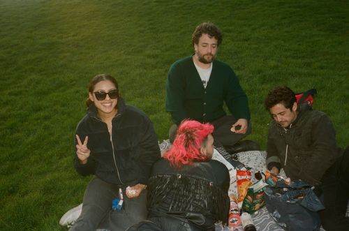 Four people enjoy a sunny picnic on a grassy field, sharing snacks and drinks in a relaxed, outdoor setting with casual attire.